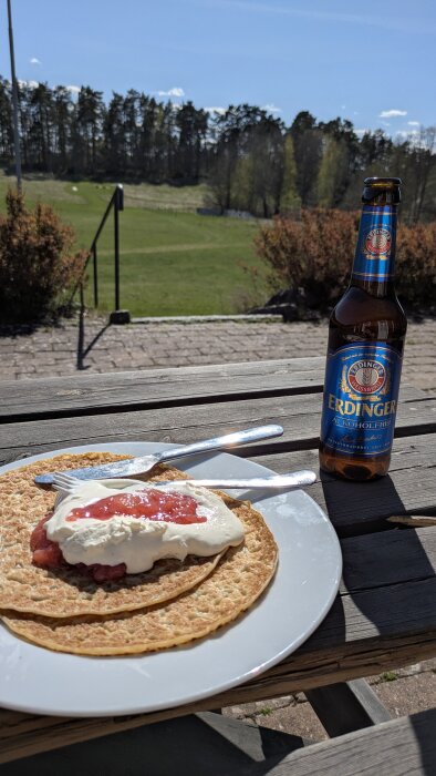 Pannkakor med sylt och grädde på tallrik, ölflaska bredvid, uteplats och grönskande landskap i bakgrunden.