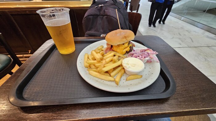 Burger med ost, pommes frites och cole slaw på en tallrik; plastglas med öl på en bricka, flygplatsmiljö i bakgrunden.