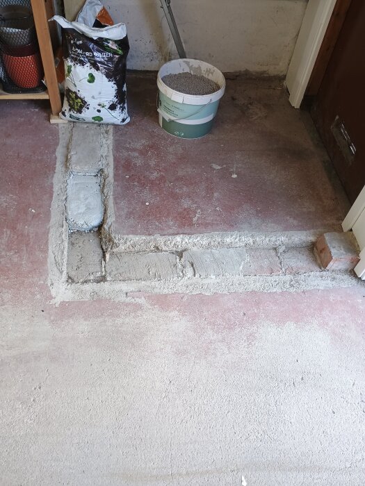 Betonglagning i garage med visad ränna, delvis fylld med betong. En nästan full säck finbetong och en hink med grus syns i bakgrunden.