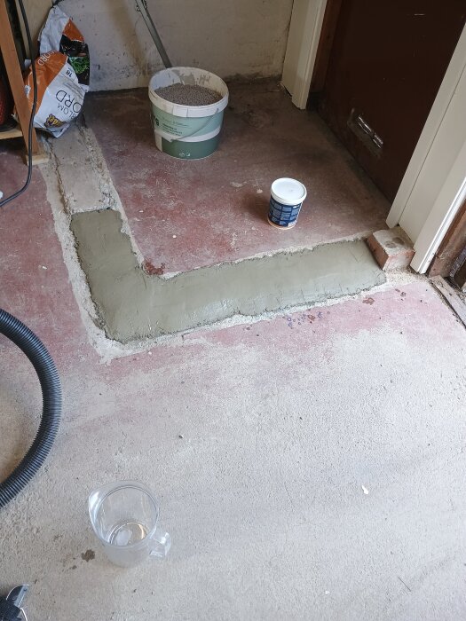Garageräffla lagad med finbetong, hink och burk syns på den rödaktiga betonggolvet. Verktyg och betongsäck i bakgrunden.