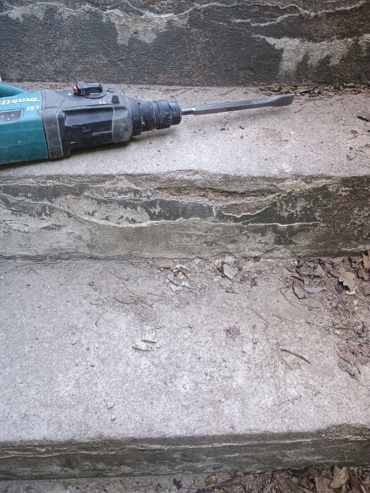 Betongtrappa med sprickor och flagnande yta, omgiven av löv och smuts. En mejsel ligger på trappsteget, redo för reparation.