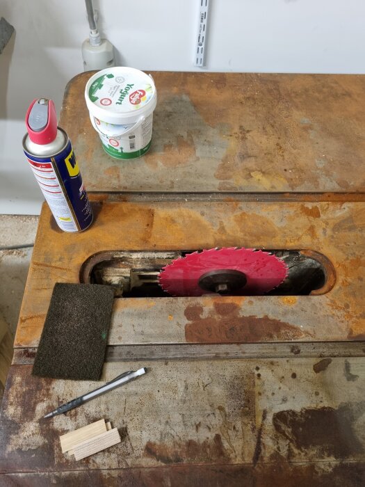 Gammal bordssåg från 60-talet med röd sågklinga, verktyg, sandpapper och WD-40. Ytrost synlig på bordet. Yogurtburk används för förvaring.