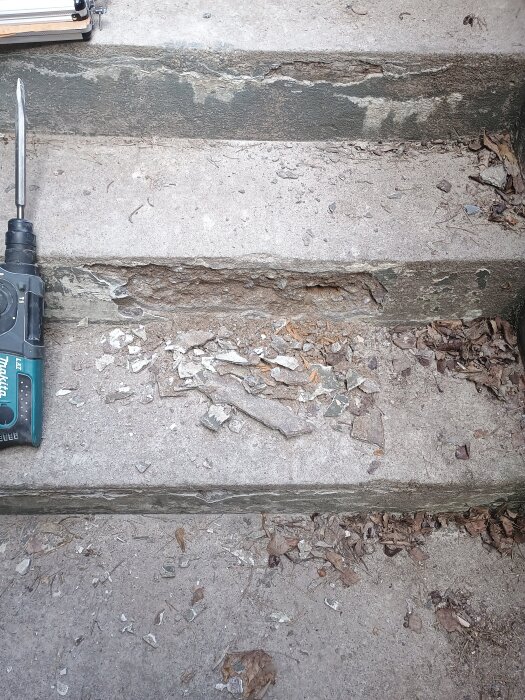 Nedgångna betongtrappsteg med sprickor och skador, omgivna av löv och byggdamm; ett verktyg ligger bredvid.