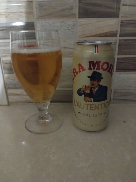 Glas med öl bredvid en Birra Moretti-burk på en ljus stenyta, med kakelvägg i bakgrunden.