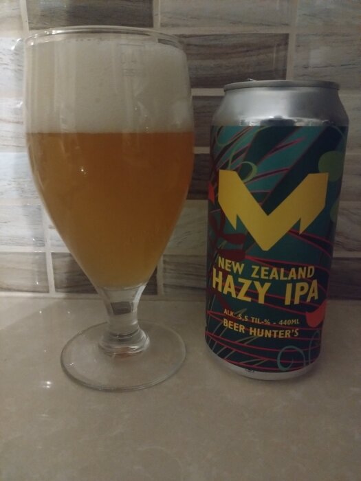 Glas med skummande öl och burk med etiketten "New Zealand Hazy IPA" från Beer Hunter's, fotograferad mot kakelvägg.