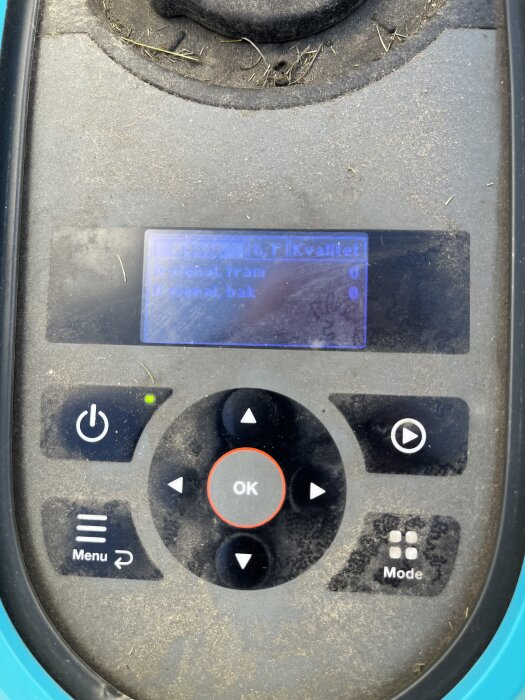 Kontrollpanel för robotgräsklippare med display som visar signal "0" och grön lampa som lyser, omgiven av smuts och gräsrester.