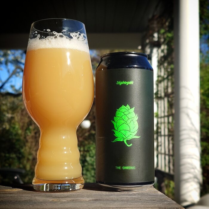 Ett glas med grumlig öl bredvid en burk med etikett föreställande humle mot en suddig bakgrund av grönska och utomhusmiljö.