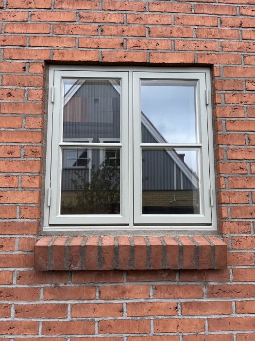 Fönster inramat i röd tegelfasad med vit karm och fyrluftsfönster, speglar en byggnad i glaset.