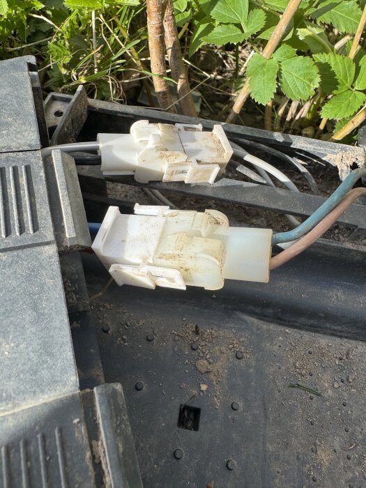 Två vita kontaktdon, delvis smutsiga och kopplade till kablar, med viss grönt lövverk i bakgrunden.