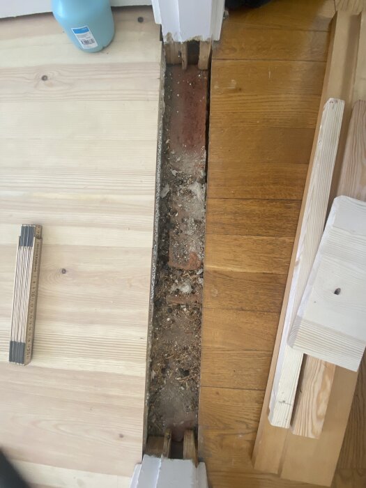 Öppen golvyta mellan trägolv där tröskeln har tagits bort, blottar ojämna tegelstenar under. Verktyg och träbitar syns vid sidan.