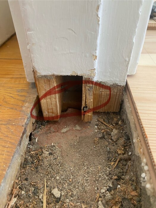 Sliten dörrkarm med borttagen tröskel, visar skador på trä och ojämn mark av tegelstenar och grus. Röda markeringar runt området där arbetet pågår.