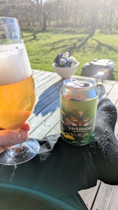 En person sitter på en altan med en burk "Vårkänslan" öl och ett glas öl i handen, med grön gräsmatta i bakgrunden.