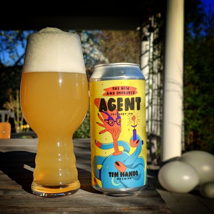 Ölglas med skummande hazy IPA bredvid en färgglad burk från "Agent" av Ten Hands Brewing på ett bord utomhus.