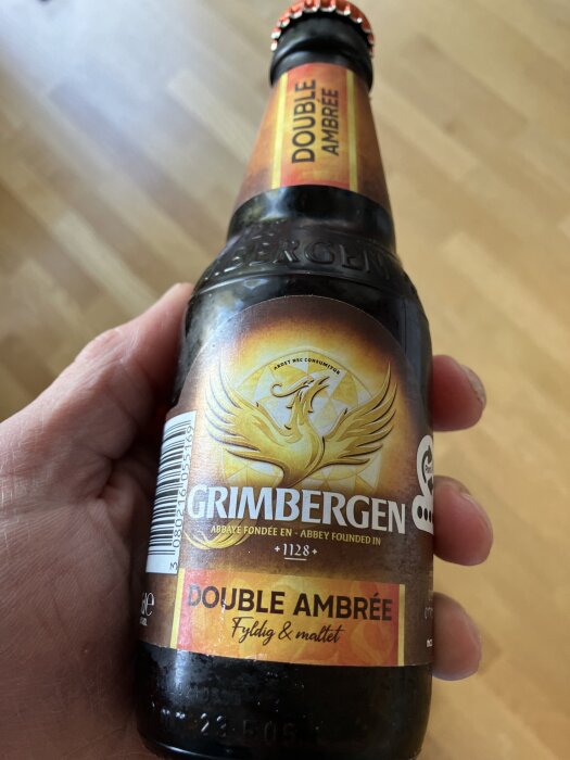 Flaska av Grimbergen Double Ambrée öl med en gyllene etikett i en hand, bakgrund av trägolv.