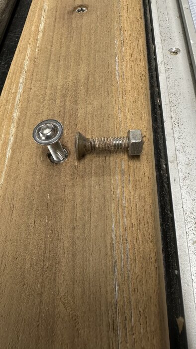Träbräda med två metallbeslag; ett fastsatt i brädan och ett löst bredvid, på ett arbetsbord.