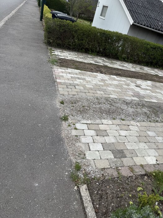 Stenlagd uppfart framför ett hus med nylagda plattor och grus, omgiven av häck och trottoar.