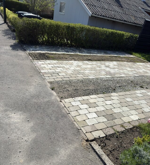 Ny stenlagd uppfart med ljusa och grå plattor mot bakgrund av en häck och hus. Grus syns mellan plattorna.