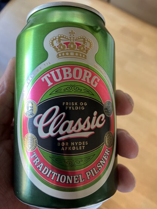 En grön ölburk med texten "Tuborg Classic" och "Traditionel Pilsner" i handen på en person, stor röd och vit etikett.