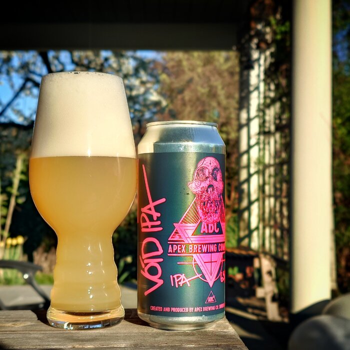Ett glas med skummig IPA och en burk med etikett som visar en dödskalle och Apex Brewing Company-logga, utomhus i solljus.