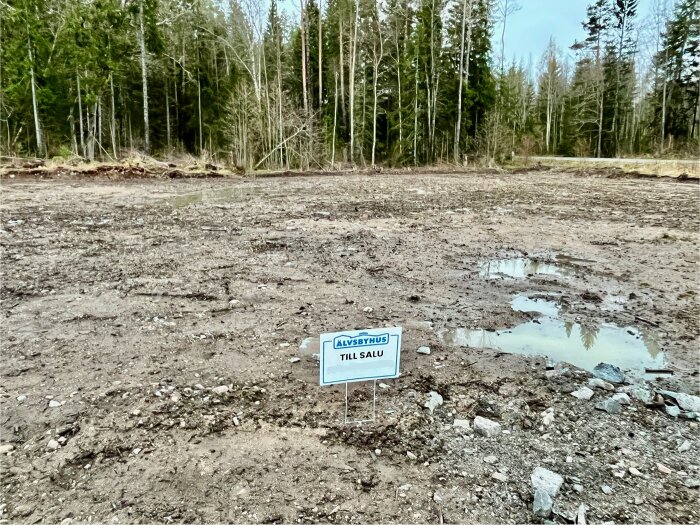 Tomt med delvis lerig mark, vattenpölar och en skylt med texten "Till Salu" placerad i mitten. Skog i bakgrunden.