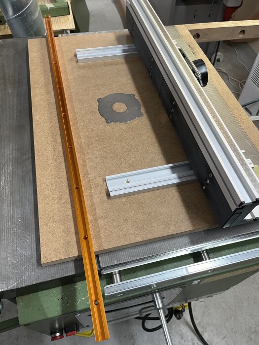 Fräsbord av MDF med skena och anhåll i orange och silver, monterat på en arbetsbänk i verkstaden.