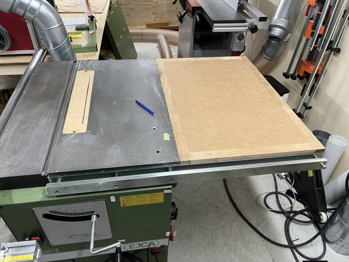 Fräsbord under konstruktion med mdf-skiva i ekram och penna placerad ovanpå, verktygsmaskiner och rörsystem synliga i bakgrunden.