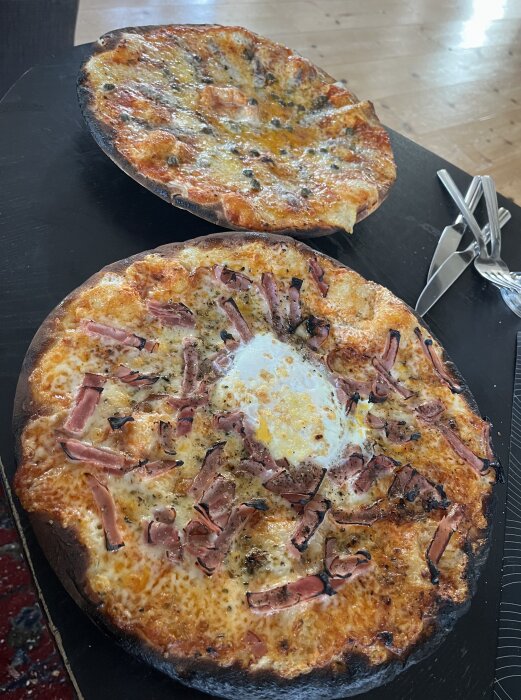 Två hembakade pizzor på svart bord; en med sardeller och kapris, den andra med skinka och ägg. Pizzakanterna är brända.