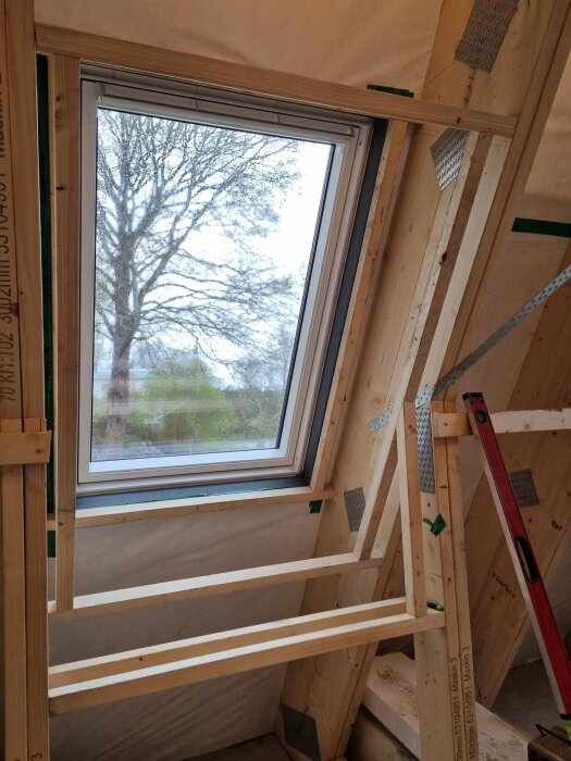 Takfönster med träram och isolering på vind, byggprojekt synligt med träreglar och vattenpass, utomhus träd syns genom fönstret.