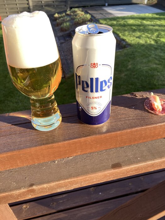 Ölglas med skum och en ölburk märkt "Pelles Filsner" på en träbalustrad i solljus, med en skiva salami bredvid.