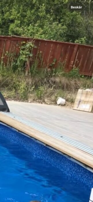 Tralldäck med pool och angränsande lerig kant och faluröd trästaket i bakgrunden.