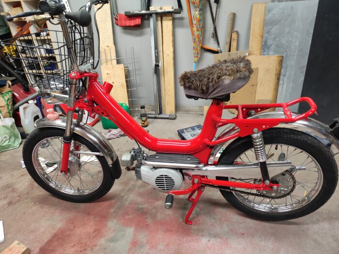 Röd moped med pälsbeklädd sadel, parkerad i ett garage med verktyg och byggmaterial i bakgrunden.