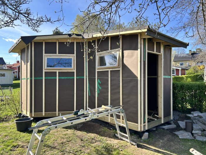 En träram med vindpapp på en byggnad, fönster och dörröppning installerade. Stege och stenar syns i trädgården bredvid.