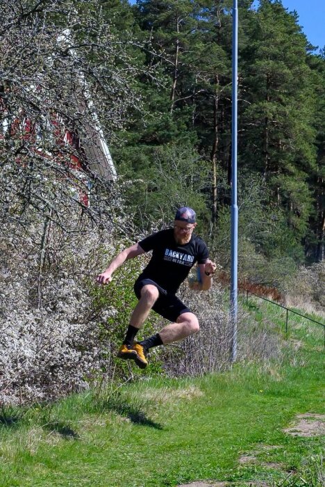 Man hoppar i luften framför grön vegetation i sportkläder och keps.
