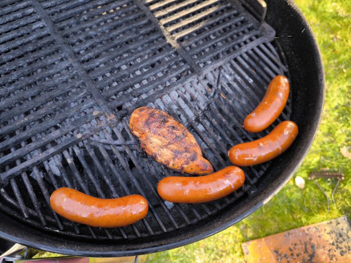 Korvar och en kycklingfilé på en grill utomhus. Grillgallret är svart med viss grillrök och kol synligt.