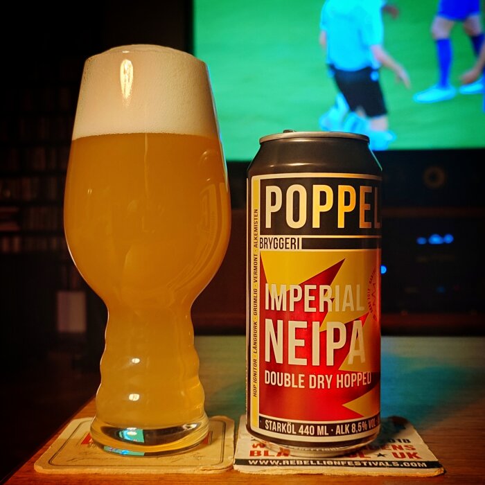 En burk Poppels Imperial NEIPA och ett fullt glas öl står på ett bord framför en pågående fotbollsmatch på TV.