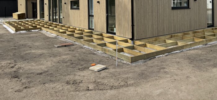 Trädäck under uppbyggnad, stomme av trä över fiberduk och cementplattor intill trähus. Stommen består av fyrkantiga sektioner, snart redo för trall.