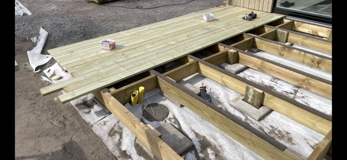 Byggprojekt pågår med delvis färdigställt trädäck av plankor på trästomme, stödd av cementplattor och fiberduk. Byggmaterial och verktyg syns på ytan.
