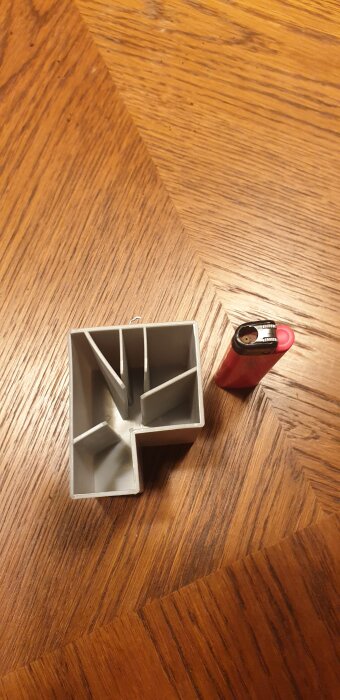 Grå plastdel med väggar i olika riktningar, placerad på träbord bredvid en röd tändare.