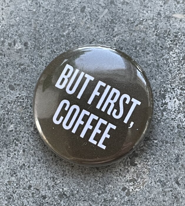 Bricka med texten "BUT FIRST, COFFEE" på en grå yta.