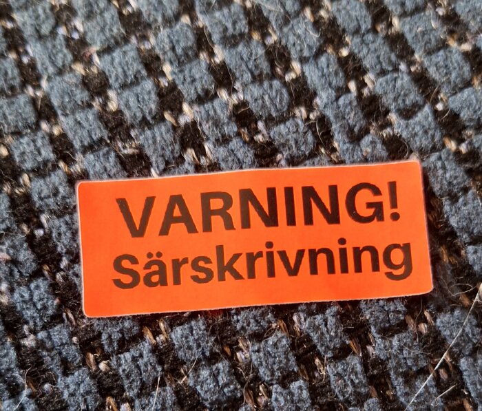 Röd etikett med texten "VARNING! Särskrivning" ligger på en mönstrad textilbakgrund.