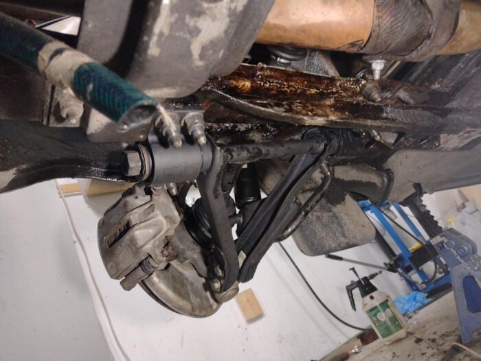 Bilens undersida med installerade nedre länkarmar och dämpare, del av bromssystemet synligt. Tecken på rostbehandling med linolja och verktyg i bakgrunden.