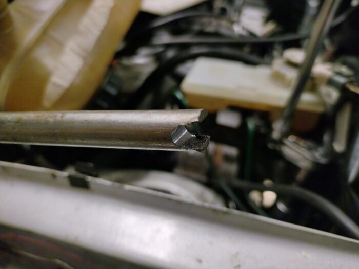 Hemgjort verktyg med låstänger för installation av stötdämpare på bilens vänstra sida, i närbild mot en bakgrund av motordelar.