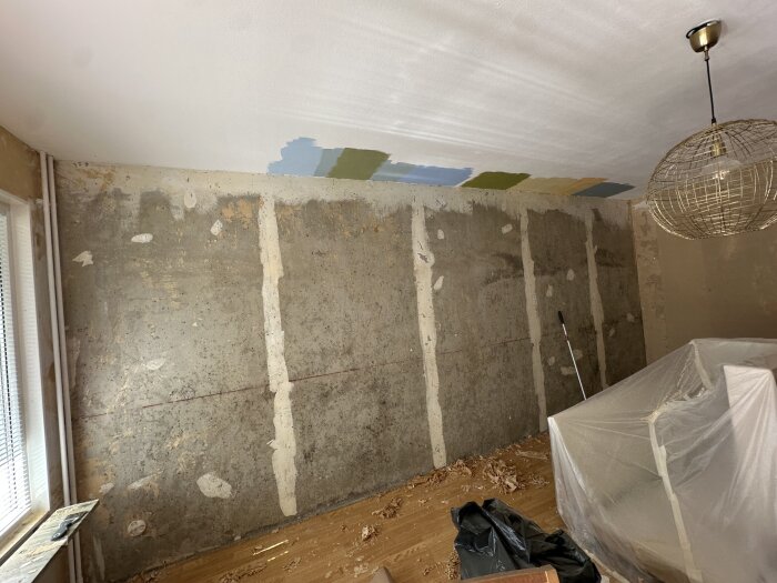 Rum med avskrapade väggar efter tapetborttagning, synliga fuktfläckar, avtäckt möbel, provmålade färger på taket, lampa och persienn.