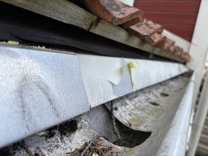 Närbild av en hängränna vid ett hus där färgen har släppt på takfotsplåten, visar skador och slitningar på ytan.