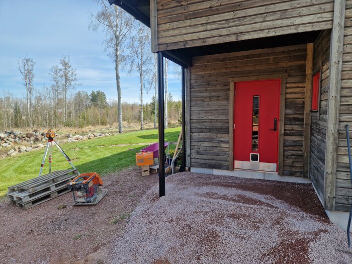 Entré med röd dörr, omgiven av grus. Verktyg och utrustning ligger på gräsmattan intill ett träskjul; skogsscen i bakgrunden.