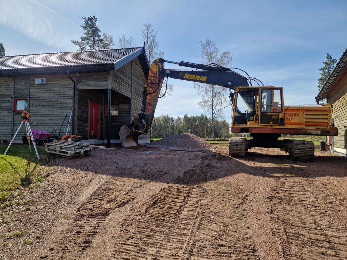 Grävmaskin vid hus på grusplan, omgiven av utrustning och pallar. Soligt väder, träden i bakgrunden. Rent och förberett för byggprojekt.