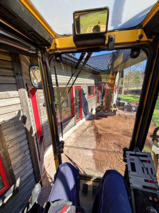 Förarens perspektiv inifrån en grävmaskin vid ett hus med röda fönster, förbereder marken för altanbygge.