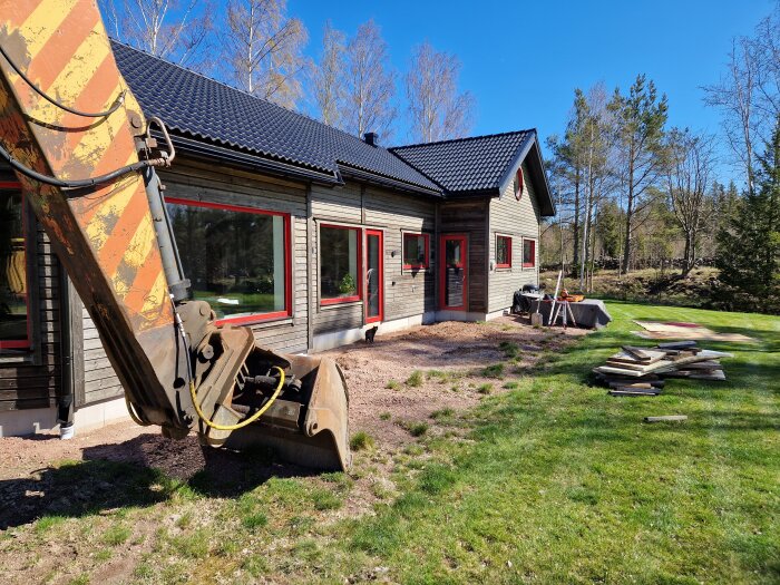 En grävmaskin vid sidan av ett trähus, omgiven av grus och gräs. Plankor och verktyg finns intill huset under en klarblå himmel.