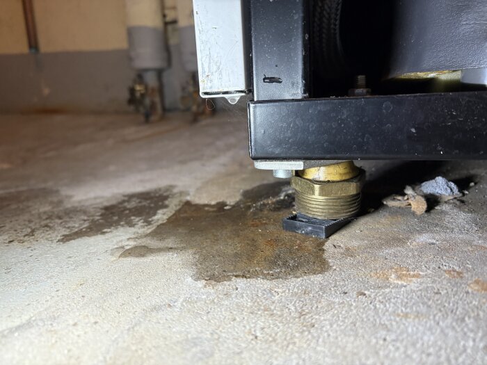 Läckande anslutning under bergvärmepump, tydlig fuktfläck på golvet, närbild av rostig metallkomponent.
