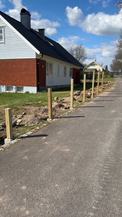 Stolpar uppställda längs en grusad väg bredvid ett hus, förberedelse för att bygga ett plank.